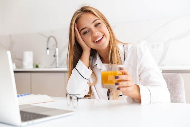femme blonde souriante en pyjama travaillant avec un ordinateur portable et buvant du jus assis à table dans une cuisine lumineuse