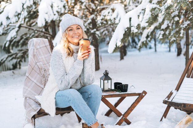 Femme blonde souriante en vêtements d'hiver avec une tasse de boisson chaude assise dans la forêt d'hiver enneigée