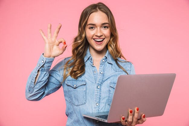 Femme blonde souriante vêtue d'une chemise en jean montrant un signe ok tout en tenant un ordinateur portable et en regardant à l'avant par-dessus le mur rose