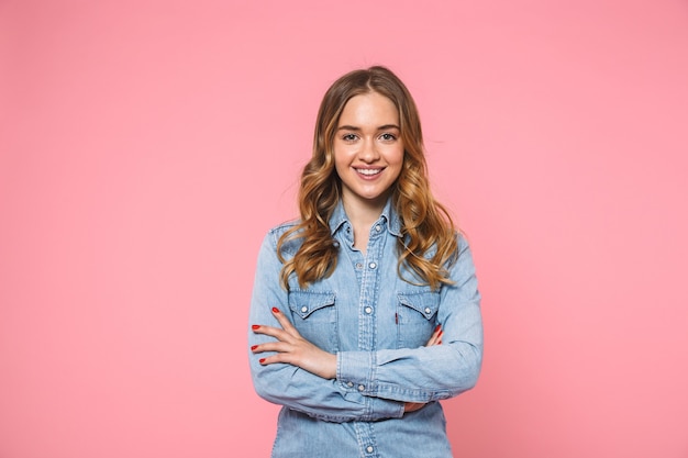 Femme blonde souriante vêtue d'une chemise en jean posant les bras croisés et regardant l'avant sur le mur rose