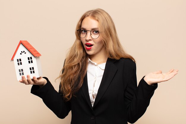 Femme blonde à la surprise et choqué, avec la mâchoire tombée tenant un objet avec une main ouverte sur le côté