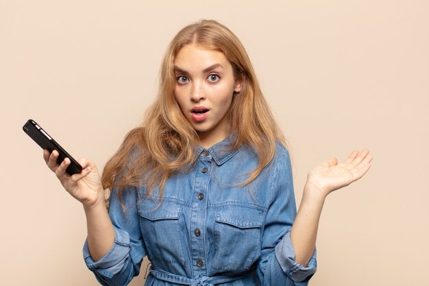 Femme blonde à la surprise et choqué, avec la mâchoire tombée tenant un objet avec une main ouverte sur le côté