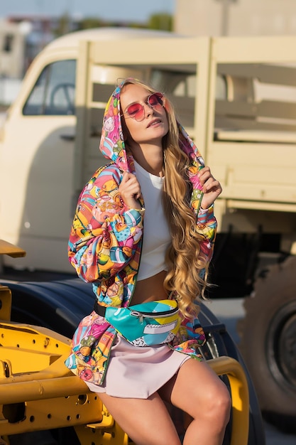 Photo femme blonde en tenue colorée à la mode ajustant le capot et regardant la caméra tout en étant assis sur le véhicule dans la rue de la ville