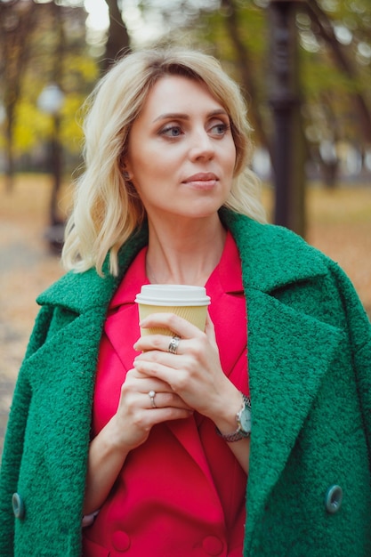 Une femme blonde tient une tasse de papier avec du café à emporter dans ses mains chaudes debout dans un parc en automne