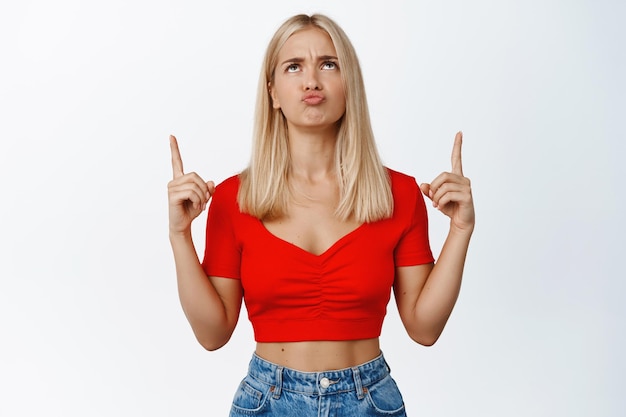 Femme blonde triste et déçue pointant et levant les yeux avec une moue bouleversée expression du visage debout dans des vêtements élégants sur fond blanc