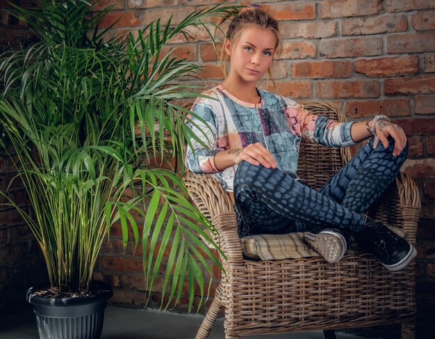 Une femme blonde urbaine est assise sur une chaise dans une pièce avec une palme verte et le mur d'une brique.