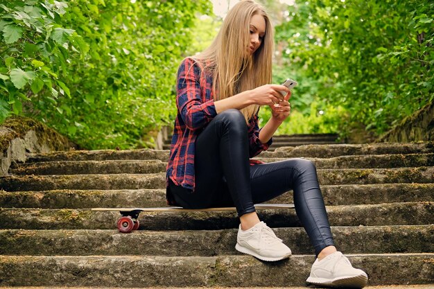 Femme blonde utilisant un smartphone et assise sur Longboard sur les marches.