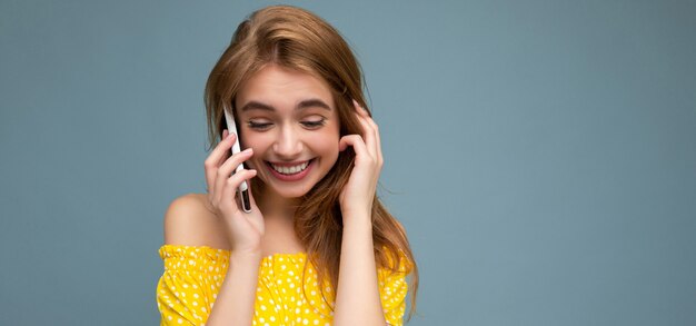 femme blonde vêtue d'une élégante robe d'été jaune debout isolé sur fond bleu tenant et parler au téléphone mobile à la recherche vers le bas.