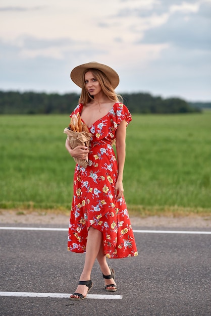 Une femme blonde vêtue d'une robe rouge et d'un chapeau de paille se tient au bord de la route