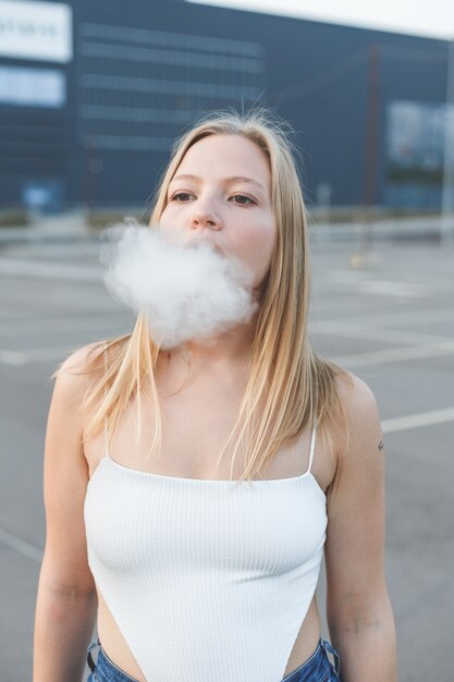 Femme blonde en ville avec vapotage de cigare électronique en t-shirt blanc à l'extérieur