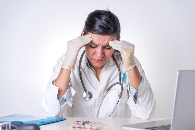 Une femme en blouse blanche se tient la tête dans la douleur et regarde un ordinateur portable médecin fatigué