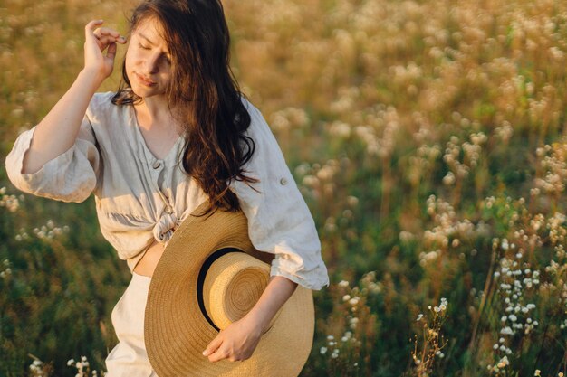 Femme bohème élégante avec un chapeau de paille posant parmi les fleurs sauvages dans la lumière chaude du coucher du soleil Délice d'été et voyage