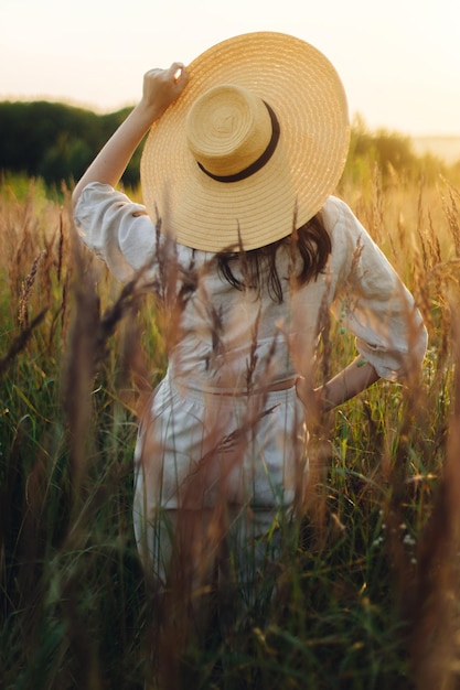 Femme bohème élégante avec un chapeau de paille posant parmi les herbes sauvages au coucher du soleil vue arrière légère Délice et tranquillité d'été Jeune femme insouciante en toile de lin rustique se relaxant dans la prairie d'été