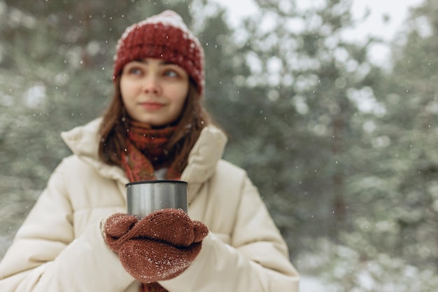 femme avec une boisson chaude dans une tasse thermos debout dans la forêt d'hiver dans les chutes de neige et les mains qui se réchauffent