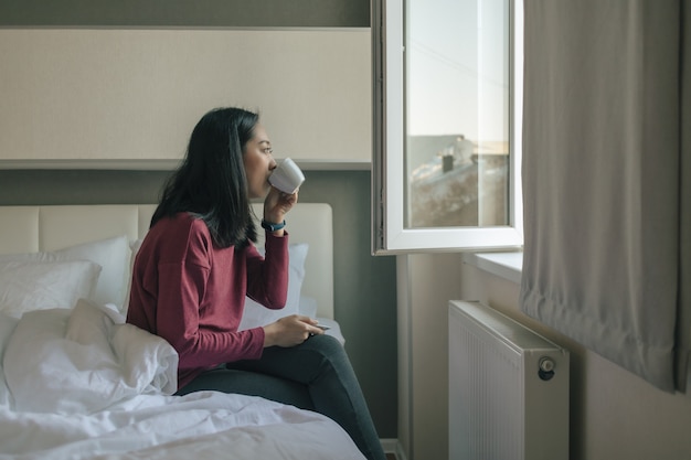 Une femme boit du café chaud et regarde par la fenêtre de son appartement.