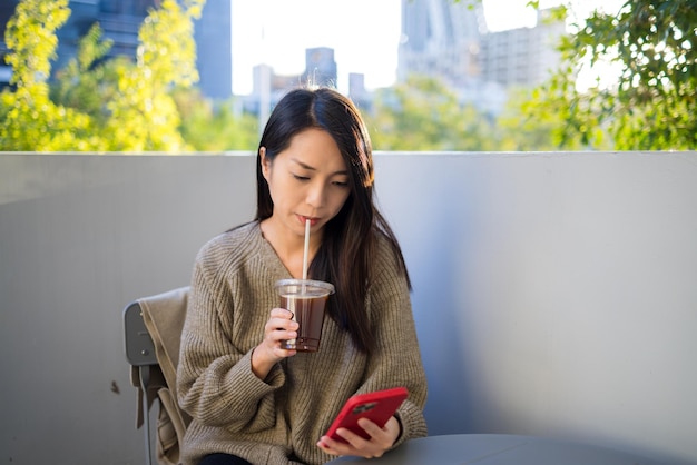 Une femme boit du café glacé en utilisant un téléphone portable dans un café en plein air