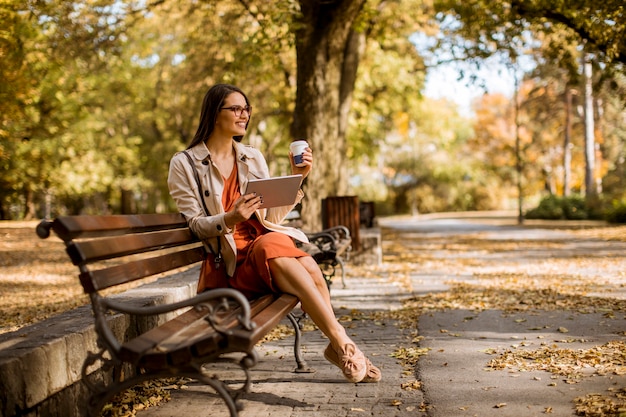 Une femme boit du café et s'assoit sur un banc dans un parc par temps d'automne, utilise une tablette et consulte les médias sociaux.