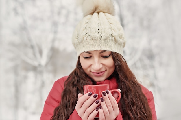 Femme boit du thé chaud ou du café de tasse à Cozy Snowy House Garden le matin d'hiver. Belle femme appréciant l'hiver à l'extérieur avec une tasse de boisson chaude. Vacances de Noël. Mode de vie d'hiver confortable.