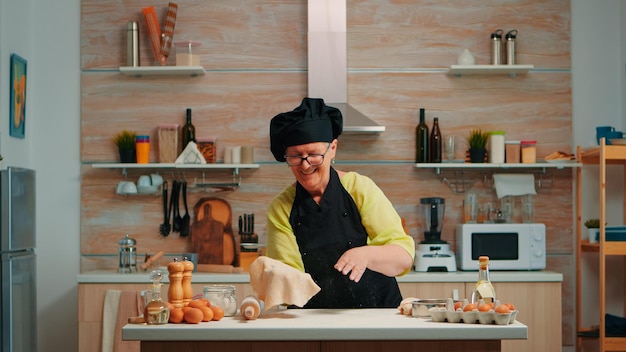 Femme avec bonete s'amusant à cuisiner à la maison dans une cuisine moderne, faisant de la poussière avec de la farine. Chef âgé à la retraite habile portant l'uniforme tournant et jetant la pâte à pizza en la jetant