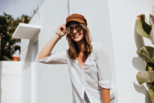 Femme de bonne humeur touche coquettement ses cheveux ondulés Dame en chemise de coton et casquette en velours côtelé sourit à côté du bâtiment dans la station balnéaire