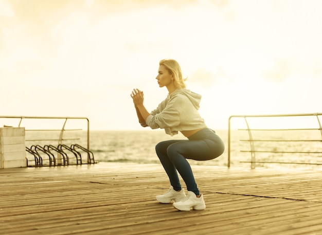 Femme en bonne santé s'entraînant sur la promenade du bord de mer. Sportive en vêtements de sport faisant des exercices de squat sur la plage au lever du soleil