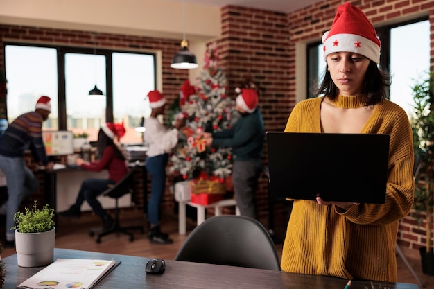 Femme en bonnet de noel utilisant un ordinateur portable dans un bureau festif, travaillant sur le lieu de travail avec un sapin de noël et des ornements. Collègues décorant l'espace de travail avec des lumières de Noël saisonnières.