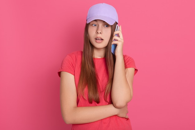 Femme avec la bouche ouverte vêtue d'une tenue décontractée parlant à quelqu'un via un téléphone intelligent moderne, ayant une expression faciale étonnée, debout avec de grands yeux isolés sur un mur de roses.