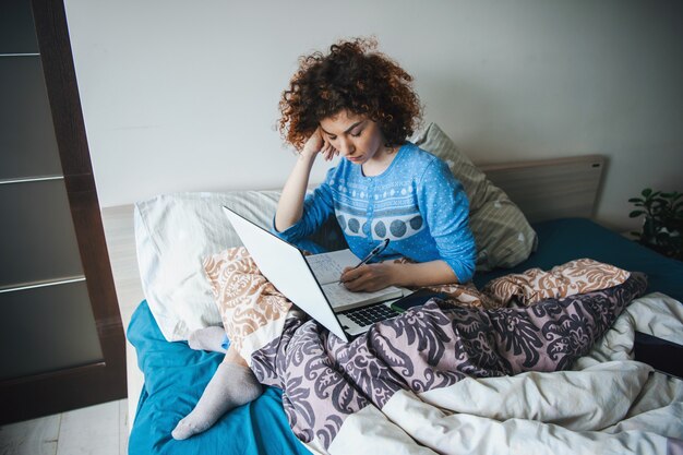 Photo femme bouclée étudiant à distance assis dans un lit en désordre mode de vie moderne de femme