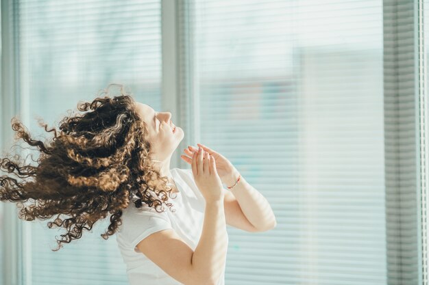 La femme bouclée heureuse écoutant la musique près de la fenêtre