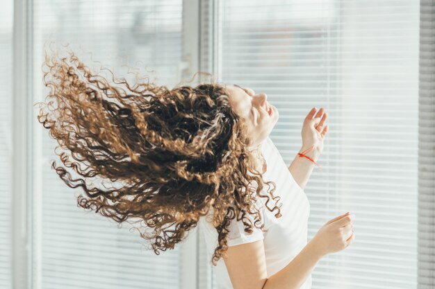 La femme bouclée heureuse écoutant la musique près de la fenêtre