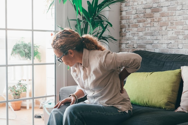 Une femme bouclée malsaine s'assoit sur un canapé à la maison qui s'étire souffrant de problèmes de dos mal surmené massage tactile féminin bas de la colonne vertébrale ayant des douleurs ou des tensions musculaires Concept de posture incorrectexA