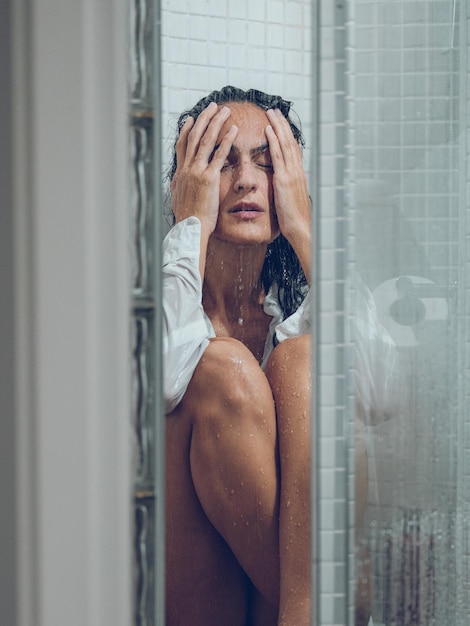 Femme bouleversée assise sur le sol de la cabine de douche sous les éclaboussures d'eau et fermant les yeux et le visage avec les mains