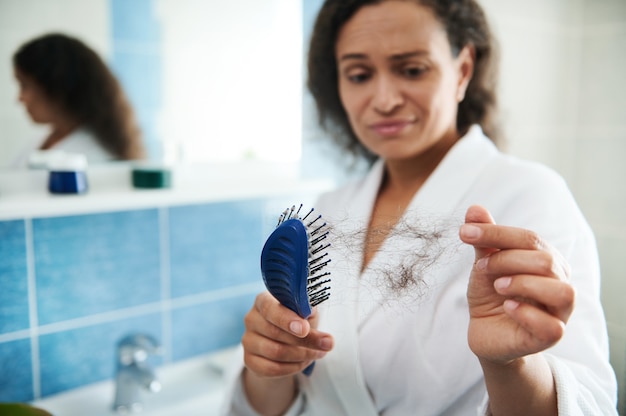 Femme bouleversée et choquée dans la salle de bain en regardant ses cheveux tomber après s'être peignée avec un peigne