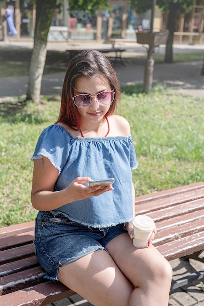 Femme branchée joyeuse aux cheveux rouges buvant du café au parc prenant selfie