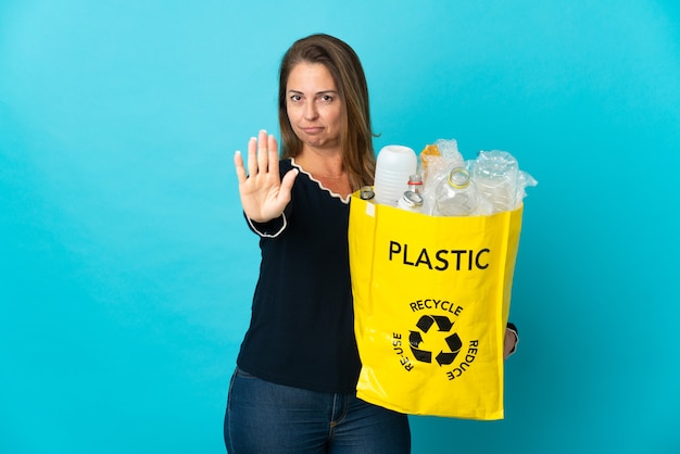 Femme brésilienne d'âge moyen tenant un sac plein de bouteilles en plastique à recycler sur bleu faisant le geste d'arrêt