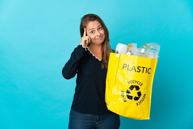 Femme brésilienne d'âge moyen tenant un sac plein de bouteilles en plastique à recycler sur bleu en pensant une idée