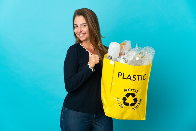 Femme brésilienne d'âge moyen tenant un sac plein de bouteilles en plastique à recycler sur le mur bleu fier et satisfait de soi