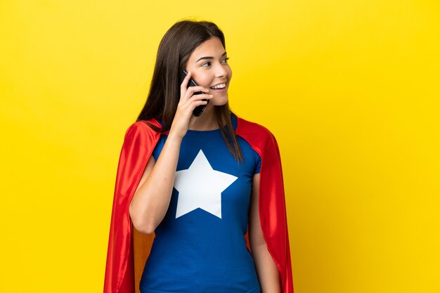 Femme brésilienne de super héros isolée sur fond jaune gardant une conversation avec le téléphone portable avec quelqu'un