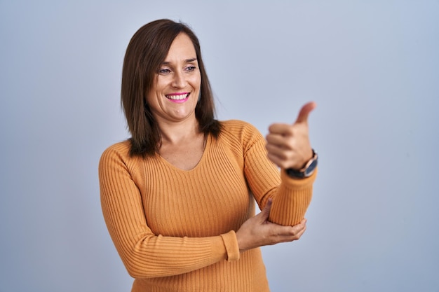 Femme brune d'âge moyen debout portant un chandail orange à la fierté souriante faisant un geste de pouce levé sur le côté