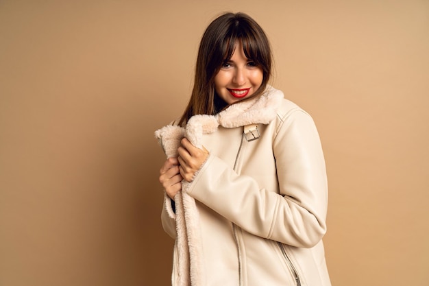 Femme brune assez élégante portant un manteau d'hiver en fourrure posant sur fond beige, maquillage tendance lumineux. Humeur positive, tenue de blogueuse.
