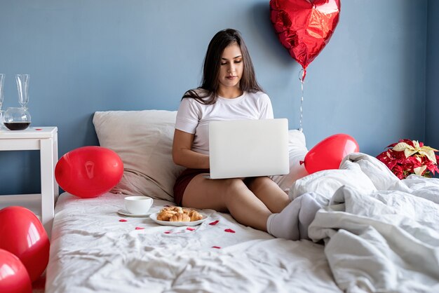 Femme brune assise dans le lit avec des ballons en forme de coeur rouge travaillant sur l'ordinateur portable