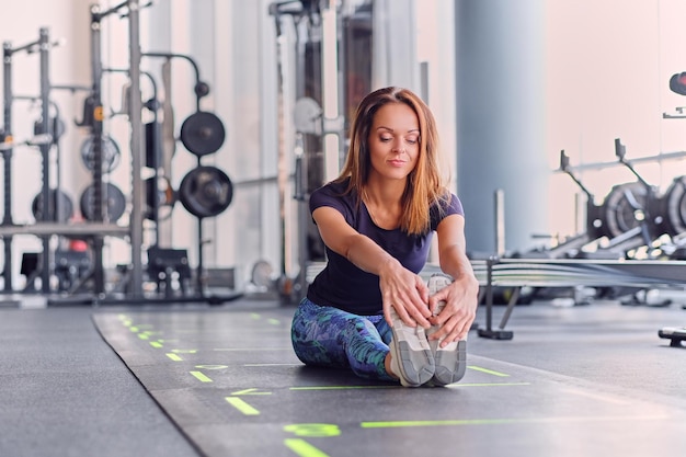 Femme brune athlétique en tenue de sport colorée qui s'étend sur un sol dans un club de gym.