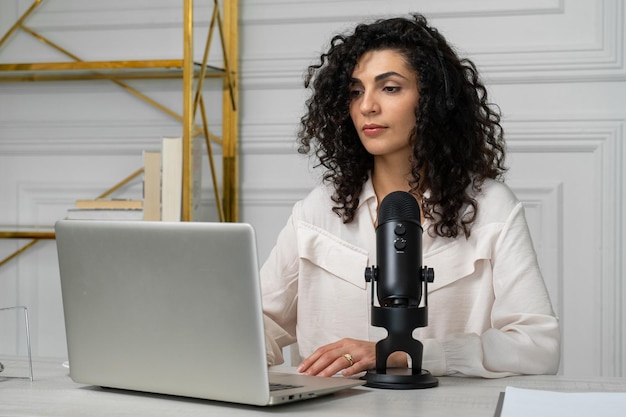 Femme brune aux cheveux bouclés avec un microphone et un ordinateur portable parlant et enregistrant un podcast dans le