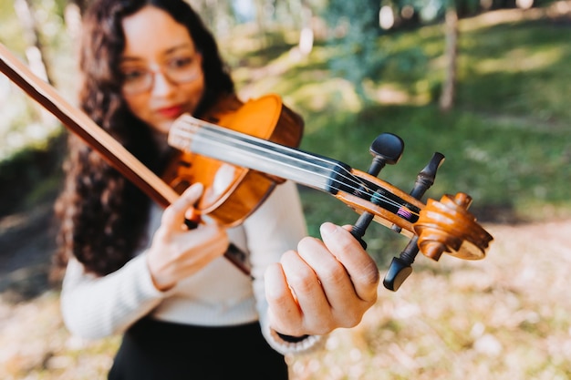 Femme brune bouclée avec des lunettes accordant son violon à l'extérieur dans les bois Espace de copie