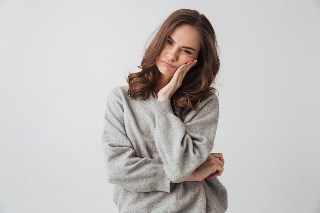 Femme brune bouleversée en pull touchant sa joue sur un mur gris