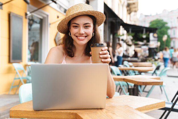 femme brune caucasienne souriante et utilisant un ordinateur portable assis dans un café d'été de rue avec du café à emporter