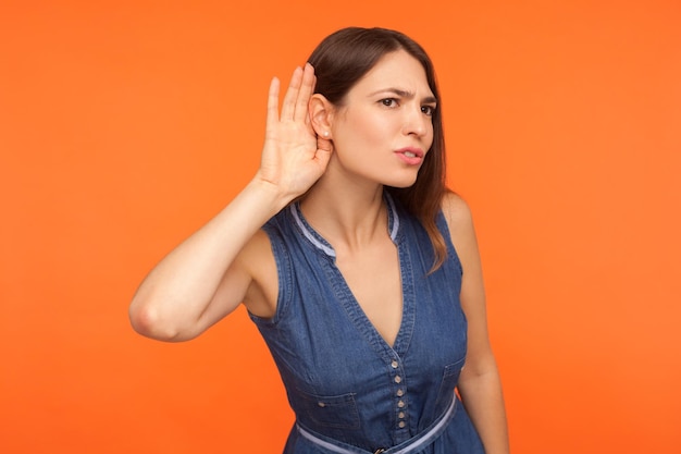 Femme brune curieuse et attentive en robe denim écoutant attentivement des informations secrètes conversation privée tenant la main près de l'oreille pour mieux entendre la prise de vue en studio intérieur isolée sur fond orange