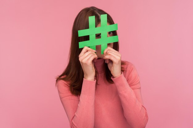 Femme brune curieuse en pull rose regardant à travers le signe hashtag vert, à la recherche de messages appropriés dans les médias sociaux, espionnage. Studio intérieur tourné isolé sur fond rose