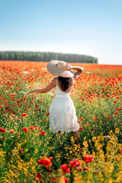 Une femme brune dans un champ de coquelicots et de marguerites Elle porte un grand chapeau de paille et une robe en dentelle blanche Vue de dos