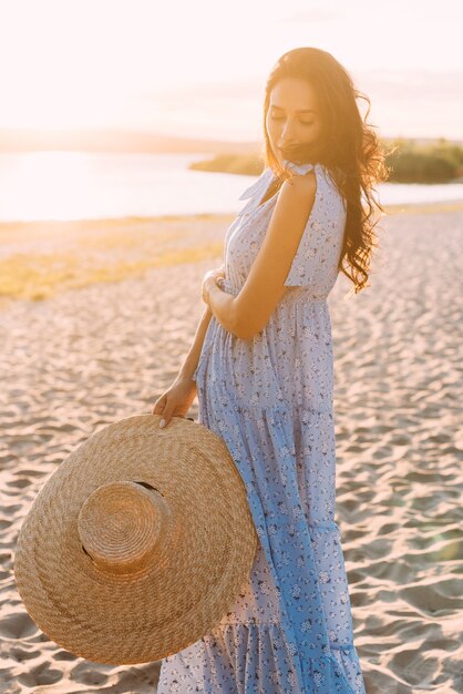 femme brune dans une élégante tenue d'été et un chapeau de paille dans les rayons du soleil sur une promenade dans la nature
